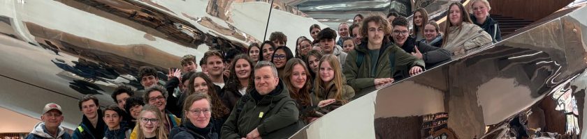 Gruppe von Menschen, die lächelnd in einem modernen, reflektierenden Raum posiert. Im Hintergrund sind gewundene, metallische Strukturen sichtbar.