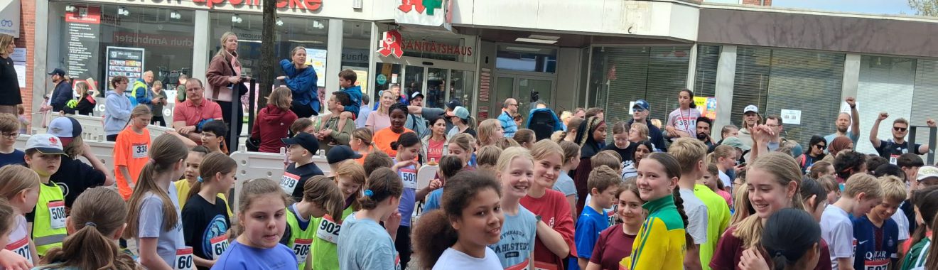 Eine große Gruppe von Kindern und Jugendlichen steht versammelt auf einer Straße, bereit für ein Rennen. Sie tragen unterschiedliche Sportbekleidung und nummerierte Startnummern. Im Hintergrund sind Gebäude und eine Baumreihe zu sehen. Es herrscht eine fröhliche und aufgeregte Atmosphäre.