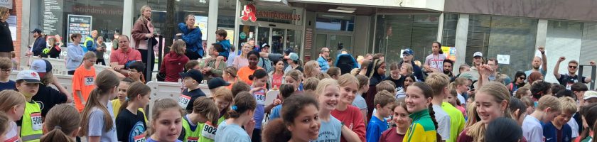Eine große Gruppe von Kindern und Jugendlichen steht versammelt auf einer Straße, bereit für ein Rennen. Sie tragen unterschiedliche Sportbekleidung und nummerierte Startnummern. Im Hintergrund sind Gebäude und eine Baumreihe zu sehen. Es herrscht eine fröhliche und aufgeregte Atmosphäre.
