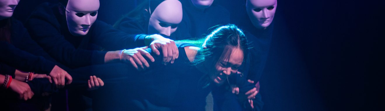 Eine Person mit langen Haaren wird von mehreren Menschen in schwarzen Anzügen und weißen Masken umgeben. Die Szene ist in schwaches, blaues Licht getaucht, wodurch eine dramatische Stimmung entsteht. Die umgebenden Personen greifen sanft nach der Hauptperson, die eine nachdenkliche oder verletzliche Haltung einnimmt.