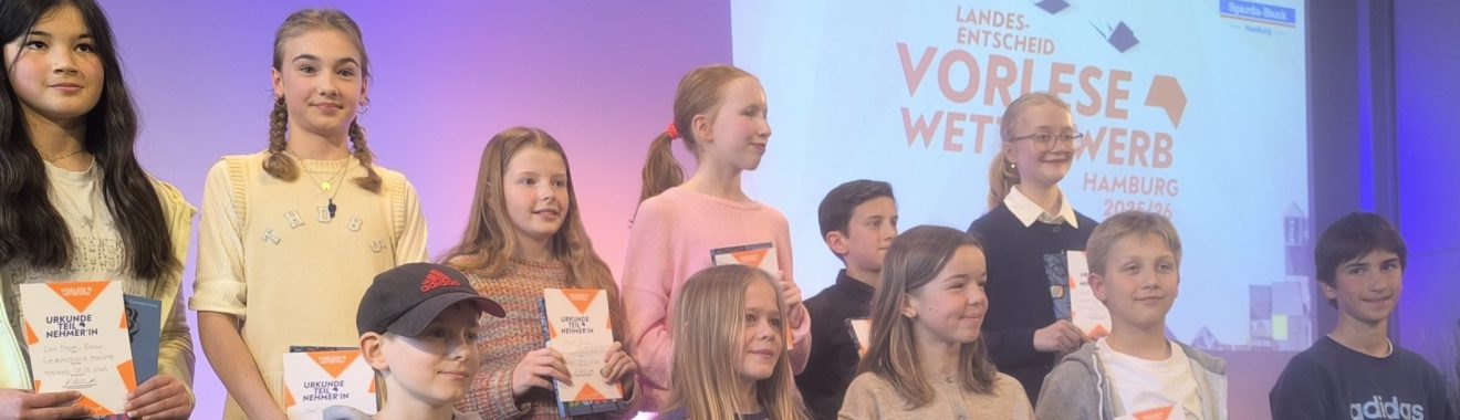 Gruppe von Kindern und Jugendlichen, die auf einer Bühne stehen. Sie lächeln und halten Urkunden in den Händen. Im Hintergrund ist ein Banner mit dem Text "Vorlese Wettbewerb" zu sehen. Die Beleuchtung ist freundlich und hebt die Teilnehmer hervor.