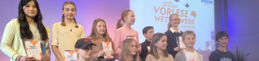 Gruppe von Kindern und Jugendlichen, die auf einer Bühne stehen. Sie lächeln und halten Urkunden in den Händen. Im Hintergrund ist ein Banner mit dem Text "Vorlese Wettbewerb" zu sehen. Die Beleuchtung ist freundlich und hebt die Teilnehmer hervor.