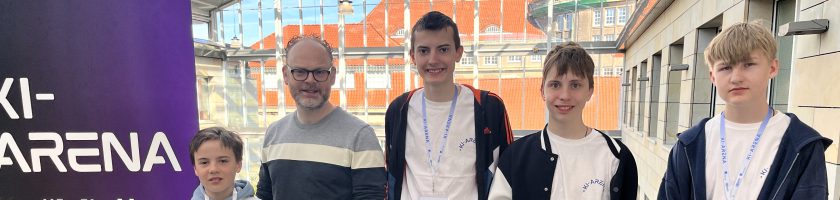 Fünf Personen stehen vor einem großen Banner mit der Aufschrift "KI-Arena". Im Hintergrund ist ein heller, offener Raum mit Fenstern zu sehen. Die Personen sind in lässiger Kleidung gekleidet, einige tragen Namensschilder. Sie wirken freundlich und stehen in einer Reihe.