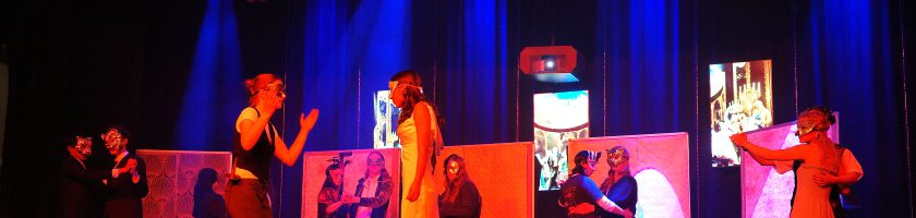 Eine Theateraufführung auf einer Bühne mit mehreren Schauspielern, die in verschiedenen Kostümen agieren. Die Bühne ist in rotes und blaues Licht getaucht, und im Hintergrund sind große Plakate oder Bilder sichtbar. Die Schauspieler interagieren miteinander, während einige das Publikum anlächeln oder applaudieren.