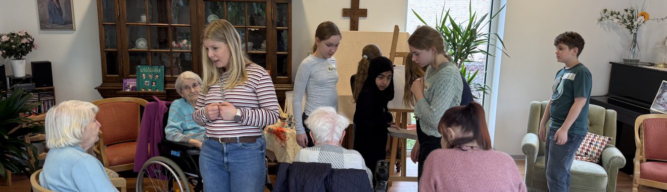 Eine Gruppe von Menschen unterschiedlichen Alters, darunter ältere Personen in Rollstühlen und jüngere Personen, versammelt sich in einem hellen Raum mit Holzfußboden. Im Hintergrund sind Möbel und Pflanzen sichtbar, und es gibt einen Schrank mit Dekorationen. Einige Personen interagieren miteinander, während andere entspannt sitzen.