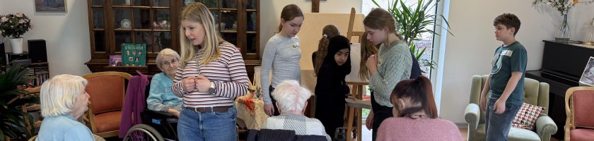 Eine Gruppe von Menschen unterschiedlichen Alters, darunter ältere Personen in Rollstühlen und jüngere Personen, versammelt sich in einem hellen Raum mit Holzfußboden. Im Hintergrund sind Möbel und Pflanzen sichtbar, und es gibt einen Schrank mit Dekorationen. Einige Personen interagieren miteinander, während andere entspannt sitzen.