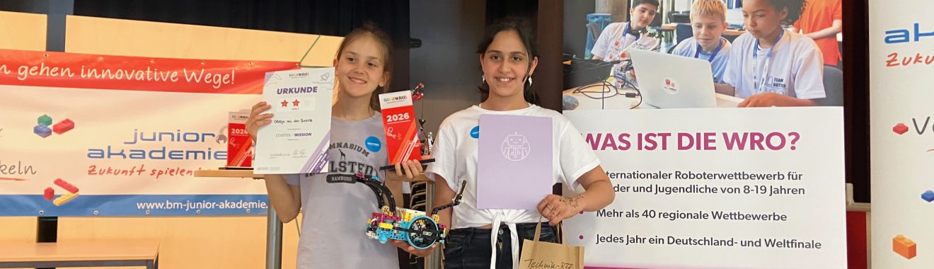 Zwei Mädchen stehen in einem Veranstaltungsraum und halten stolz ihre Auszeichnungen in den Händen. Sie tragen T-Shirts und lächeln in die Kamera. Im Hintergrund sind Banner mit Informationen zur WRO (World Robot Olympiad) sowie zur Junior Akademie zu sehen. Auf einem Tisch sind weitere Materialien platziert.