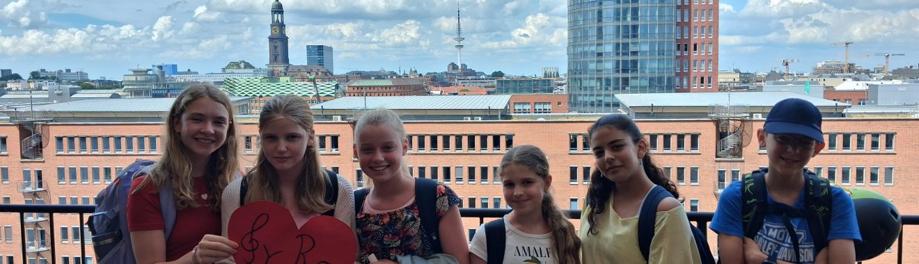 Sechs Kinder stehen auf einem Balkon mit Blick auf eine Stadtlandschaft. Sie halten ein rotes Herz in der Mitte der Gruppe. Im Hintergrund sind moderne Gebäude und ein blauer Himmel mit einigen Wolken sichtbar. Die Kinder tragen sommerliche Kleidung und wirken fröhlich.