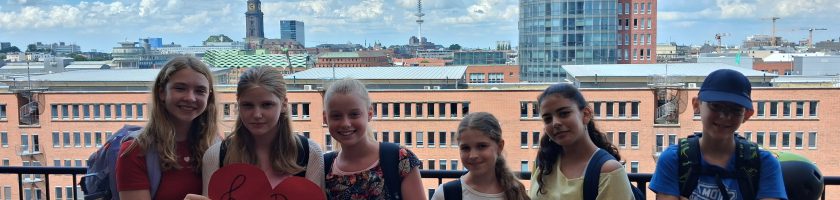 Sechs Kinder stehen auf einem Balkon mit Blick auf eine Stadtlandschaft. Sie halten ein rotes Herz in der Mitte der Gruppe. Im Hintergrund sind moderne Gebäude und ein blauer Himmel mit einigen Wolken sichtbar. Die Kinder tragen sommerliche Kleidung und wirken fröhlich.