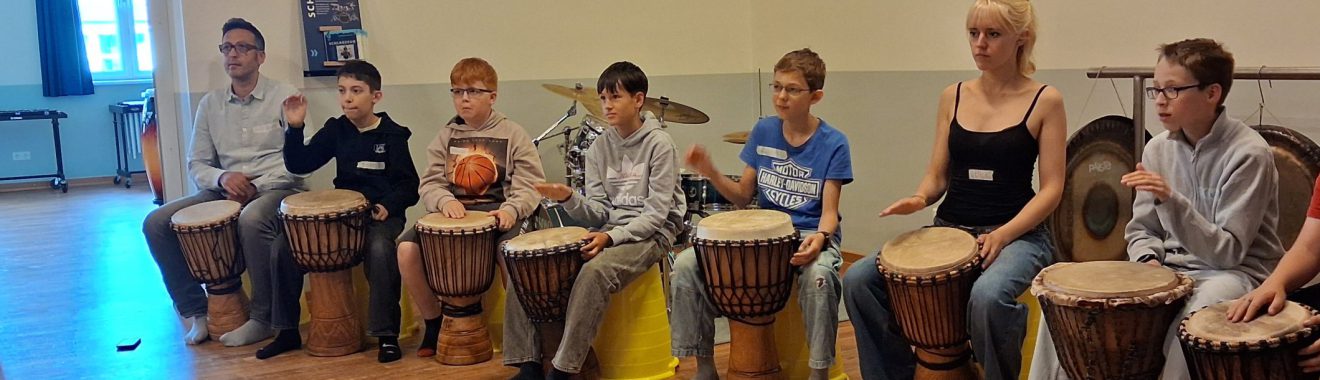 Eine Gruppe von Kindern und Jugendlichen sitzt an Trommeln in einem Raum. Sie spielen gemeinsam und konzentriert, einige schauen auf die Trommeln, während andere den Takt halten. Die Atmosphäre wirkt kreativ und gemeinschaftlich.