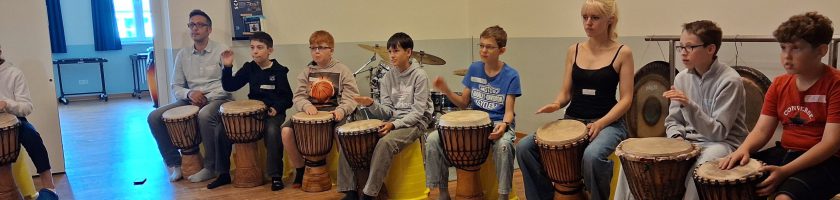 Eine Gruppe von Kindern und Jugendlichen sitzt an Trommeln in einem Raum. Sie spielen gemeinsam und konzentriert, einige schauen auf die Trommeln, während andere den Takt halten. Die Atmosphäre wirkt kreativ und gemeinschaftlich.
