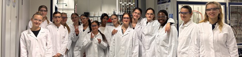 Gruppe von 15 Personen in weißen Laborkitteln, die in einem Labor stehen und lächeln. Einige halten ihre Hände in die Höhe oder umarmen sich. Im Hintergrund sind Laborausrüstungen und Regale sichtbar.