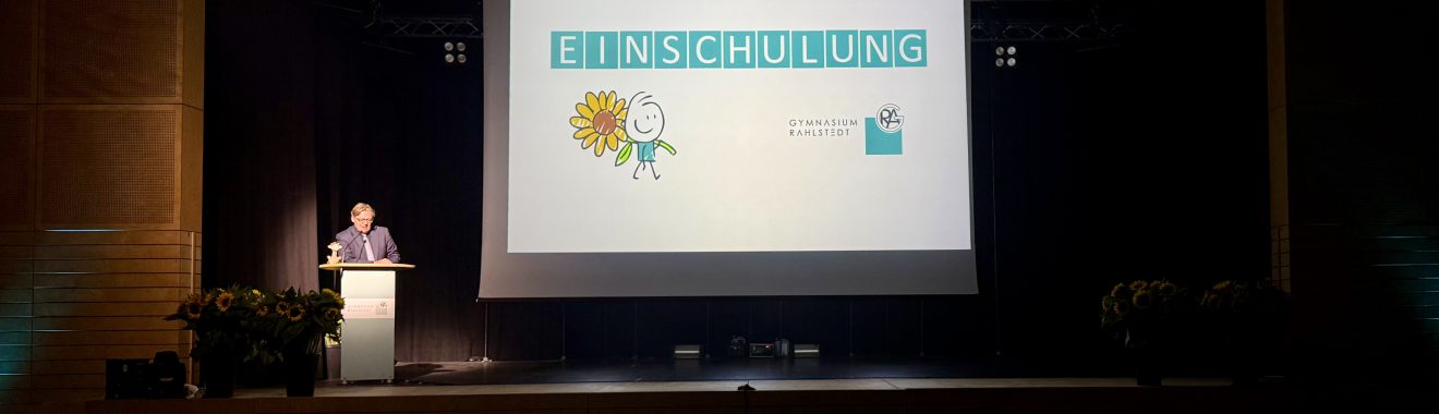 Ein Mann steht an einem Rednerpult auf einer Bühne. Im Hintergrund ist eine Präsentation mit dem großen Wort „EINSCHULUNG“ und grafischen Elementen, darunter eine Sonne und ein Schulranzen. Der Raum ist mit Pflanzen und einer Publikumssilhouette ausgestattet.
