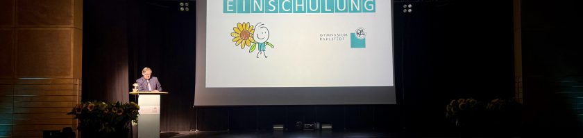 Ein Mann steht an einem Rednerpult auf einer Bühne. Im Hintergrund ist eine Präsentation mit dem großen Wort „EINSCHULUNG“ und grafischen Elementen, darunter eine Sonne und ein Schulranzen. Der Raum ist mit Pflanzen und einer Publikumssilhouette ausgestattet.