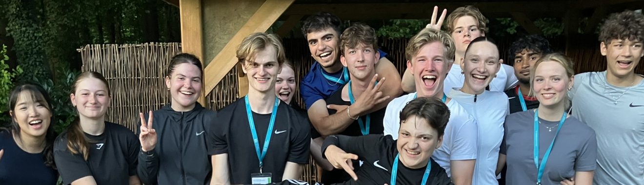 Gruppe von Jugendlichen und jungen Erwachsenen, die lächelnd und fröhlich vor einer Holzstruktur stehen. Einige machen Peace-Zeichen oder posieren lustig. Die Szene vermittelt Freude und Gemeinschaft.