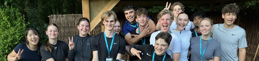 Gruppe von Jugendlichen und jungen Erwachsenen, die lächelnd und fröhlich vor einer Holzstruktur stehen. Einige machen Peace-Zeichen oder posieren lustig. Die Szene vermittelt Freude und Gemeinschaft.