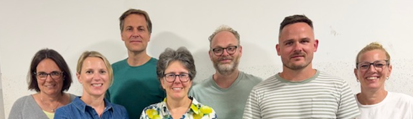 Gruppe von sieben Personen, die vor einer hellen Wand stehen. Sie lächeln und schauen in die Kamera. Die Personen tragen unterschiedliche Kleidung, darunter ein gestreiftes Shirt und ein farbenfrohes, blumiges Hemd.