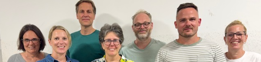 Gruppe von sieben Personen, die vor einer hellen Wand stehen. Sie lächeln und schauen in die Kamera. Die Personen tragen unterschiedliche Kleidung, darunter ein gestreiftes Shirt und ein farbenfrohes, blumiges Hemd.