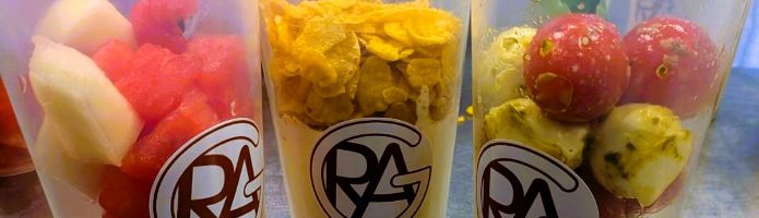 Drei durchsichtige Becher mit verschiedenen frischen Snacks stehen auf einem Tisch. Der linke Becher enthält eine Mischung aus Wassermelone und anderen Früchten, der mittlere Becher hat eine gelbe, cremige Mischung, und der rechte Becher ist gefüllt mit kleinen, runden Snacks, die grün und rot sind. Im Hintergrund sind weitere Becher mit Obst sichtbar.