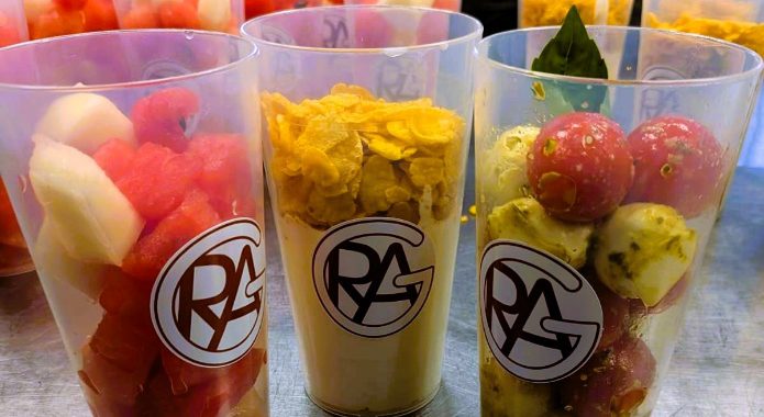 Drei durchsichtige Becher mit verschiedenen frischen Snacks stehen auf einem Tisch. Der linke Becher enthält eine Mischung aus Wassermelone und anderen Früchten, der mittlere Becher hat eine gelbe, cremige Mischung, und der rechte Becher ist gefüllt mit kleinen, runden Snacks, die grün und rot sind. Im Hintergrund sind weitere Becher mit Obst sichtbar.