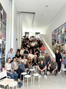 Eine große Gruppe von Menschen sitzt und steht auf einer Treppe in einem modernen Raum mit weißen Wänden. Im Hintergrund sind farbige Poster oder Bilder an der Wand zu sehen. Die Personen sind unterschiedlich gekleidet und scheinen an einer Veranstaltung oder einem Treffen teilzunehmen.