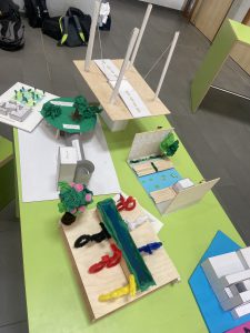 Modellbau-Projekte auf einem Tisch, darunter verschiedene Miniaturlandschaften und Strukturen aus Holz und Bastelmaterialien. Auf dem Tisch sind Bäume, Gebäude und Straßen dargestellt, die kreative architektonische Entwürfe zeigen. Die Modelle sind farbenfroh und variieren in der Größe, um verschiedene Elemente einer Stadt oder Landschaft darzustellen.