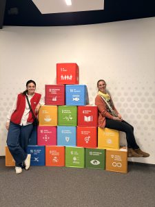 Zwei Personen sitzen auf einer bunten Stapel von Kisten, die verschiedene Symbole und Texte zeigen. Die Kisten sind in leuchtenden Farben gehalten und repräsentieren vermutlich verschiedene Themen oder Ziele. Im Hintergrund ist eine helle Wand mit einem subtilen Muster. Die Personen tragen legere Kleidung und lächeln in die Kamera.
