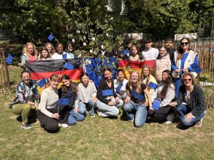 Gruppe von jungen Menschen, die auf einer Wiese stehen und lächeln. Sie halten Flaggen der Europäischen Union, der Ukraine und Deutschlands. Im Hintergrund sind Bäume und eine Blumenpracht zu sehen. Die Teilnehmer sind in verschiedenen Outfits gekleidet und zeigen ein Gefühl der Gemeinschaft und Freude.