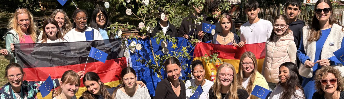 Gruppe von jungen Menschen, die auf einer Wiese stehen und lächeln. Sie halten Flaggen der Europäischen Union, der Ukraine und Deutschlands. Im Hintergrund sind Bäume und eine Blumenpracht zu sehen. Die Teilnehmer sind in verschiedenen Outfits gekleidet und zeigen ein Gefühl der Gemeinschaft und Freude.