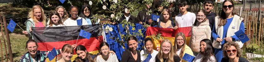 Gruppe von jungen Menschen, die auf einer Wiese stehen und lächeln. Sie halten Flaggen der Europäischen Union, der Ukraine und Deutschlands. Im Hintergrund sind Bäume und eine Blumenpracht zu sehen. Die Teilnehmer sind in verschiedenen Outfits gekleidet und zeigen ein Gefühl der Gemeinschaft und Freude.