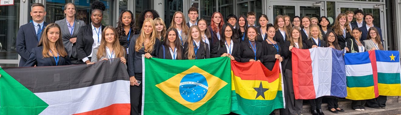 Gruppe von Menschen, die vor einem Gebäude steht und verschiedene Nationalflaggen hält, darunter die Flaggen von Sudan, Brasilien, Ghana, der Schweiz und weiteren Ländern. Die Personen tragen formelle Kleidung und lächeln in die Kamera.
