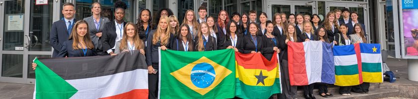 Gruppe von Menschen, die vor einem Gebäude steht und verschiedene Nationalflaggen hält, darunter die Flaggen von Sudan, Brasilien, Ghana, der Schweiz und weiteren Ländern. Die Personen tragen formelle Kleidung und lächeln in die Kamera.