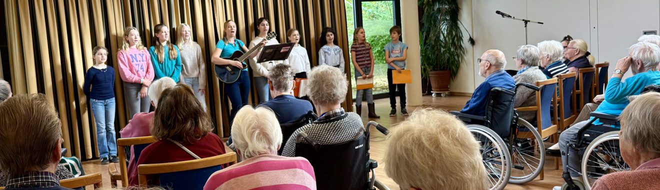 Eine Gruppe von Kindern steht auf einer Bühne und führt ein Programm auf, während eine ältere Zuhörerschaft, darunter viele Personen im Rollstuhl, aufmerksam zuschaut. Im Hintergrund sind Pflanzen und Mikrofone sichtbar.