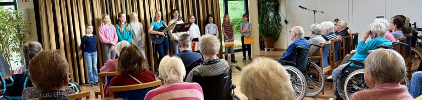 Eine Gruppe von Kindern steht auf einer Bühne und führt ein Programm auf, während eine ältere Zuhörerschaft, darunter viele Personen im Rollstuhl, aufmerksam zuschaut. Im Hintergrund sind Pflanzen und Mikrofone sichtbar.